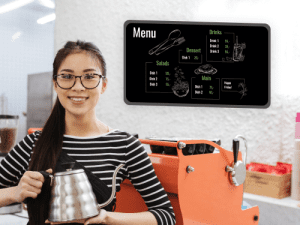 Woman holding a pot of coffe, and a digital sign in the bacground with the menu and prices.