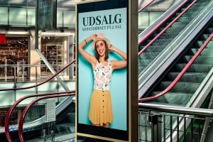 Vertical digital sign hanging by the ecalators in a mall, displaying an ad.